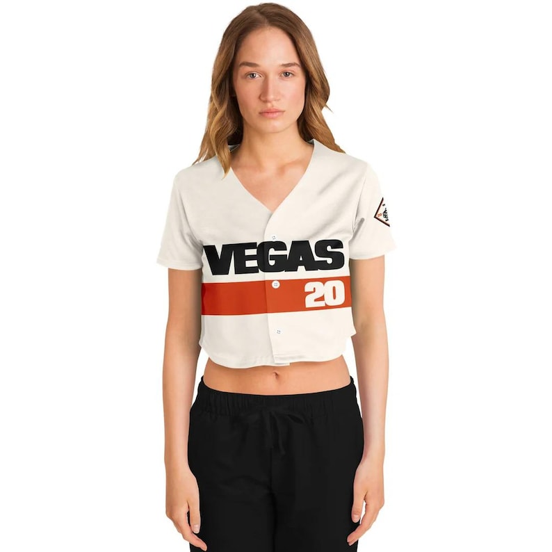 Op de afbeelding: Cr&egrave;mekleurig, gecropt honkbalshirt met het woord "VEGAS" in zwarte blokletters boven een rode streep en het nummer "20". De trui heeft een V-hals en korte mouwen. Een klein embleem staat op de rechter mouw.
