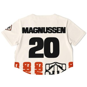 Op de afbeelding: Cr&egrave;mekleurig honkbalshirt met de naam "MAGNUSSEN" in zwarte blokletters boven het nummer "20". Het nummer is ook zwart. De onderkant van het shirt heeft het woord "GRAN" in rode letters en een zwarte afbeelding.