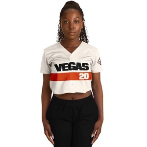 Op de afbeelding: Cr&egrave;mekleurig, gecropt honkbalshirt met het woord "VEGAS" in zwarte blokletters en het nummer "20" in rood. Een horizontale oranje streep loopt over de borst. Het shirt heeft een V-hals en korte mouwen.