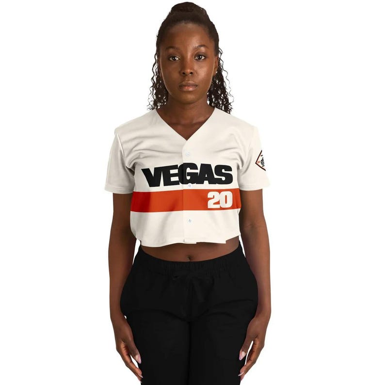 Op de afbeelding: Cr&egrave;mekleurig, gecropt honkbalshirt met het woord "VEGAS" in zwarte blokletters en het nummer "20" in rood. Een horizontale oranje streep loopt over de borst. Het shirt heeft een V-hals en korte mouwen.