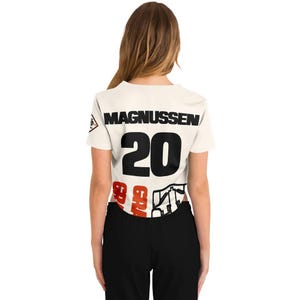 Op de afbeelding: Cr&egrave;me crop top met de naam "MAGNUSSEN" in zwarte blokletters boven het nummer "20" in zwart en rood. De onderkant van de top heeft een rood-wit grafisch ontwerp. De top wordt gedragen met een zwarte broek.