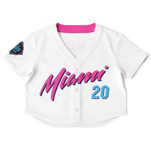 Peut inclure: Maillot de baseball blanc &agrave; col en V et manches courtes. Le maillot porte le mot "Miami" en rose avec une ombre bleue et le num&eacute;ro "20" en bleu. Un &eacute;cusson sur la manche gauche indique "The 05 Miami". Le col est rose.