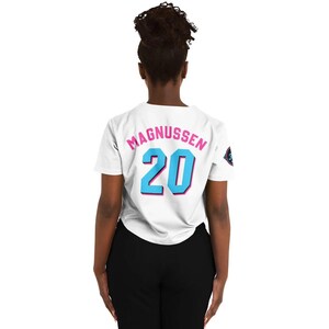 Peut inclure: T-shirt blanc avec le nom "MAGNUSSEN" en rose et le num&eacute;ro "20" en bleu avec un contour rose. Le t-shirt a un petit embl&egrave;me sur la manche droite. La personne porte un pantalon noir.