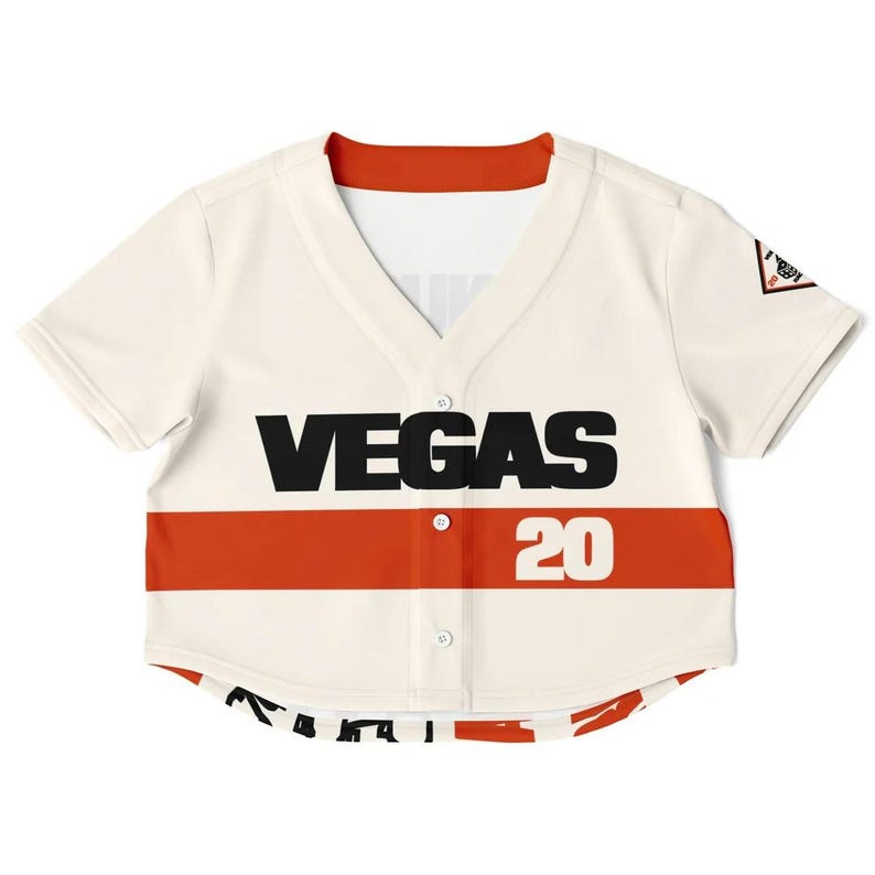Op de afbeelding: Cr&egrave;mekleurig honkbalshirt met een rode streep over de borst. Het woord "VEGAS" is gedrukt in grote zwarte letters, met het nummer "20" in wit. Het shirt heeft een V-hals en korte mouwen.