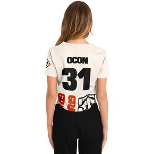 May include: Cream-colored crop top with the name "OCON" above the number "31" in black. The number is large and bold. The bottom of the shirt has red and white graphics. The person is wearing black pants.