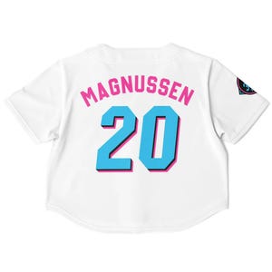 Peut inclure: Maillot de baseball blanc avec le nom "MAGNUSSEN" en lettres roses au-dessus du num&eacute;ro "20" en bleu avec un contour rose. Un logo est visible sur la manche droite. Le maillot a des manches courtes.