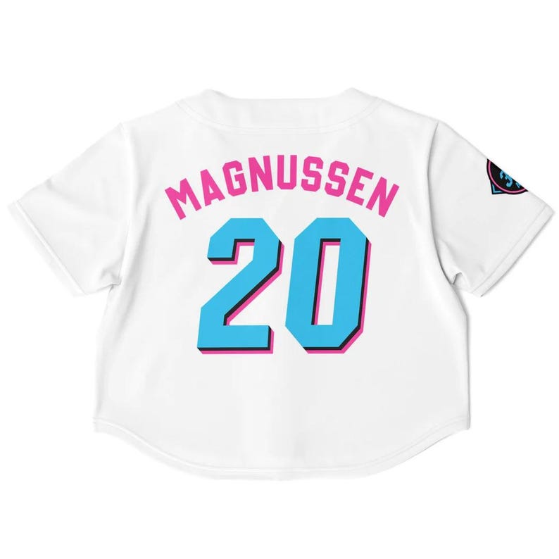Peut inclure: Maillot de baseball blanc avec le nom "MAGNUSSEN" en lettres roses au-dessus du num&eacute;ro "20" en bleu avec un contour rose. Un logo est visible sur la manche droite. Le maillot a des manches courtes.