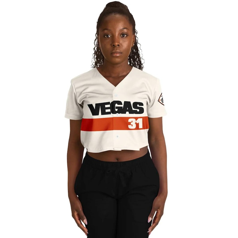 May include: Cream-colored cropped baseball jersey with a red stripe across the chest. The word "VEGAS" is printed in black above the number "31". The jersey has a V-neck and short sleeves.