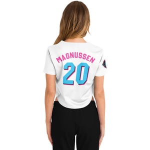 Peut inclure: Haut court blanc avec le nom "MAGNUSSEN" en rose et le num&eacute;ro "20" en bleu avec un contour rose. Un petit embl&egrave;me est sur l'&eacute;paule droite. Le haut est port&eacute; avec un pantalon noir.