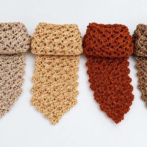 May include: Six neckties arranged in a row, each featuring an openwork crochet pattern. The ties are presented in a spectrum of colors including white, light beige, tan, rust orange, dark brown, and cream, all with a pointed bottom edge.