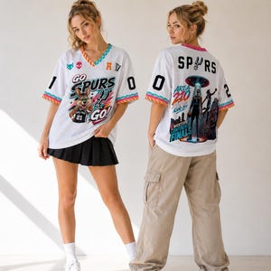 Op de afbeelding: Wit shirt in jersey-stijl met een kleurrijke afbeelding van een cartoonkonijn en de woorden "GO SPURS GO!" op de voorkant. De achterkant van het shirt toont "SPURS" en een afbeelding van een basketballer. Het shirt heeft een V-hals en kleurrijke afwerking.