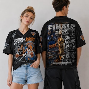 K&ouml;nnte beinhalten: Ein schwarzes T-Shirt mit V-Ausschnitt, das auf der Vorderseite eine Grafik des Basketball-Spiels Spurs gegen Knicks und auf der R&uuml;ckseite eine gro&szlig;e Grafik der Finals 2026 zeigt, inklusive Larry O'Brien Troph&auml;e, Skylines und Teamlogos.