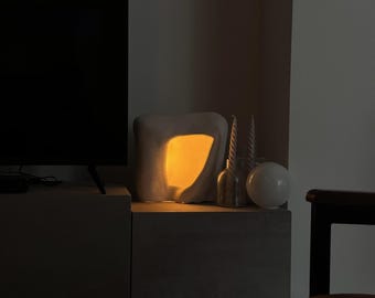 May include: A modern, abstract lamp with a warm, yellow glow emanating from within. The lamp is placed on a gray cabinet, alongside decorative glass bottles and a white sphere. The scene is dimly lit, creating a cozy atmosphere.
