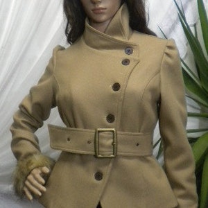 Può includere: Un cappotto color beige con collo alto e vita con cintura. Il cappotto presenta una disposizione di bottoni unica e polsini in pelliccia. La cintura ha una fibbia color oro. Il cappotto è esposto su un manichino.