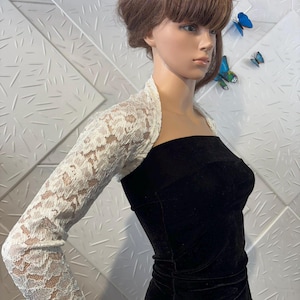 May include: A white lace bolero with long sleeves. The bolero is worn over a black velvet dress. The lace has a floral pattern. The mannequin is positioned in front of a textured white wall with blue butterfly decorations.