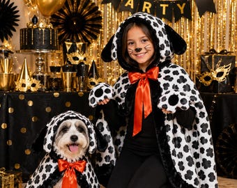 May include: A matching set of two dalmatian-inspired capes featuring white fabric with black spots, black faux fur trim, and a bright red satin ribbon bow at the neck. Each cape includes an attached hood with floppy black ears and paw-shaped hand covers.