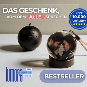 Könnte beinhalten: Ein schwarzer kugelförmiger Lautsprecher und eine transparente Glaskugel mit einem Foto eines Paares darin. Die Kugel steht auf einem dunklen Sockel und hat ein beiges und schwarzes Schlüsselband. Auf dem Bild steht "DAS GESCHENK, VOM DEM ALLE SPRECHEN" und "BESTSELLER".