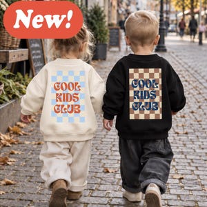Könnte beinhalten: Zwei langärmelige Sweatshirts, eines in Creme und eines in Schwarz, mit einem rechteckigen Aufdruck auf der Rückseite, der ein Karomuster und den Text "COOL KIDS CLUB" in Wellenschrift zeigt. Das cremefarbene Sweatshirt hat ein blau-weißes Karomuster, das schwarze ein braun-weißes.