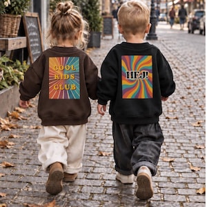 Könnte beinhalten: Ein braunes Sweatshirt mit einer rechteckigen Grafik mit farbigem Strahlenmuster und dem Text COOL KIDS CLUB sowie ein schwarzes Sweatshirt mit einer rechteckigen Grafik mit farbigem Wirbelmuster und dem Text HEJ!.