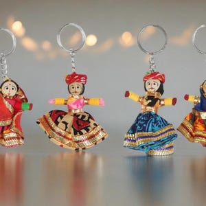 May include: Four handmade doll keychains. Each doll has a painted face, colorful clothing, and a silver keychain ring. The dolls are dressed in traditional Indian attire, with vibrant colors and gold trim.
