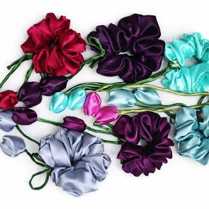 May include: A collection of satin scrunchies in shades of red, purple, teal, pink, and grey. The scrunchies are arranged to look like flowers, with green stems and leaves, forming a decorative and colourful arrangement.