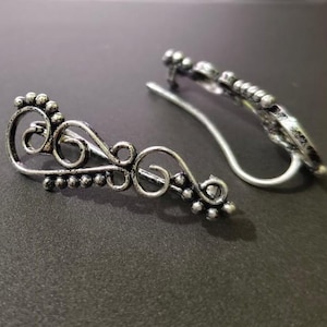 Puede incluir: Dos puños de oreja plateados con un diseño ornamental y arremolinado. Cada puño presenta una serie de pequeñas cuentas redondas y líneas curvas delicadas. Los puños de oreja se muestran sobre una superficie oscura.