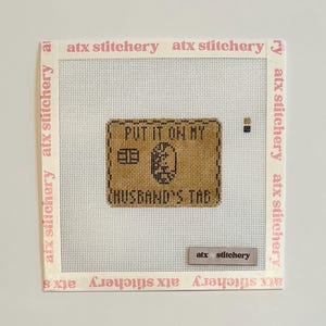 Puede incluir: Un bordado a punto de cruz con el texto "PUT IT ON MY" encima de "HUSBAND'S TAG" en hilo negro sobre un fondo marr&oacute;n. Las palabras "atx stitchery" est&aacute;n impresas en rosa en los bordes.