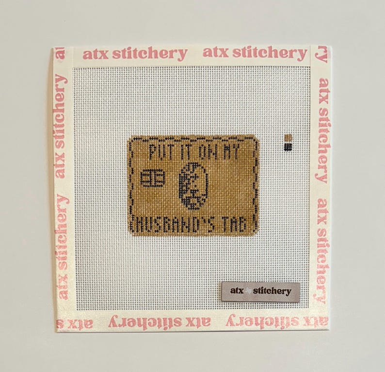 Puede incluir: Un bordado a punto de cruz con el texto "PUT IT ON MY" encima de "HUSBAND'S TAG" en hilo negro sobre un fondo marr&oacute;n. Las palabras "atx stitchery" est&aacute;n impresas en rosa en los bordes.