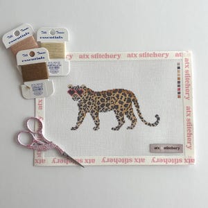 May include: A needlepoint canvas featuring a leopard wearing sunglasses, stitched in shades of brown, tan, and black. The canvas is bordered with the text "atx stitchery." Three spools of thread in beige and brown tones and a pair of pink scissors are also present.