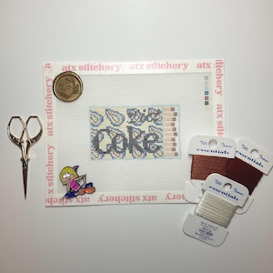 May include: A needlepoint project with the word "Coke" stitched in black and blue, surrounded by paisley designs. Gold scissors, thread spools, and a pin are arranged around the canvas. The canvas is on a white surface.
