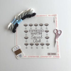May include: A needlepoint kit with the text "Espresso Martini Social Club" surrounded by martini glasses. The kit includes white, gray, and black embroidery floss, a spool of brown thread, and pink scissors. The canvas is 12 inches by 12 inches.