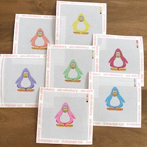 May include: A collection of seven cross-stitch canvases featuring colorful penguin designs. Each penguin is a different color, including yellow, pink, purple, green, and blue, with orange feet and a white belly. The canvases have a white border with the text "atx stitchery".