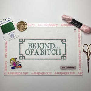 May include: A needlepoint canvas with the text "BE KIND... OF A BITCH" in dark green. The canvas is surrounded by pink trim with the words "atx stitchery". Accessories include green thread, pink thread, a gold pin, and small gold scissors.
