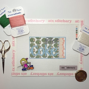 May include: A needlepoint project with the word "Lake" stitched in blue. The project is surrounded by spools of thread in green, pink, and white, along with gold scissors, a pin, and a wax seal. The background is white.