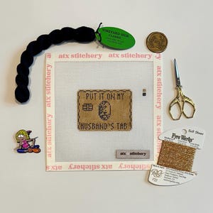 Puede incluir: Un conjunto de suministros de punto de aguja, que incluye un lienzo con "PUT IT ON MY HUSBAND'S TAB" bordado, hilo negro, tijeras doradas y una bobina de hilo dorado. Tambi&eacute;n hay un alfiler y una moneda.