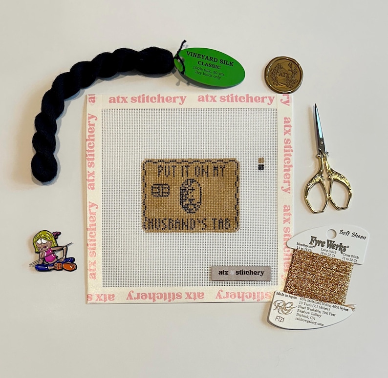Puede incluir: Un conjunto de suministros de punto de aguja, que incluye un lienzo con "PUT IT ON MY HUSBAND'S TAB" bordado, hilo negro, tijeras doradas y una bobina de hilo dorado. Tambi&eacute;n hay un alfiler y una moneda.