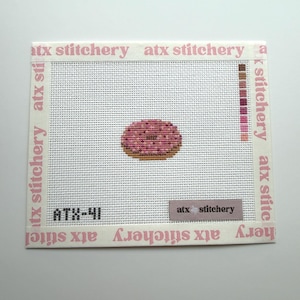 May include: A square needlepoint canvas featuring a centered pink donut with brown sprinkles. The canvas is framed by white tape printed with the text atx stitchery. A color palette guide is on the right, and the code ATX-41 is in the bottom left.