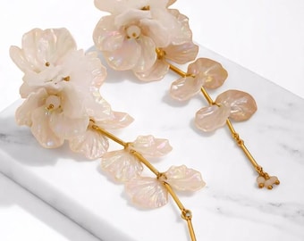 May include: A pair of floral earrings with a pearlescent finish. The earrings feature large, layered flower designs and smaller petal accents, all connected by gold-colored stems. Small, round beads dangle from the bottom of each earring.