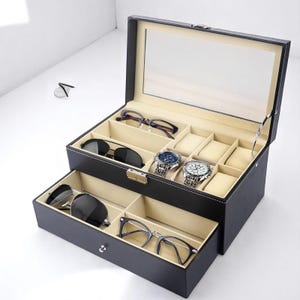 Puede incluir: Un organizador de relojes y gafas de polipiel negra con tapa de cristal transparente. El organizador tiene dos niveles, con compartimentos para relojes, gafas de sol y gafas. El interior es beige y el exterior es negro.