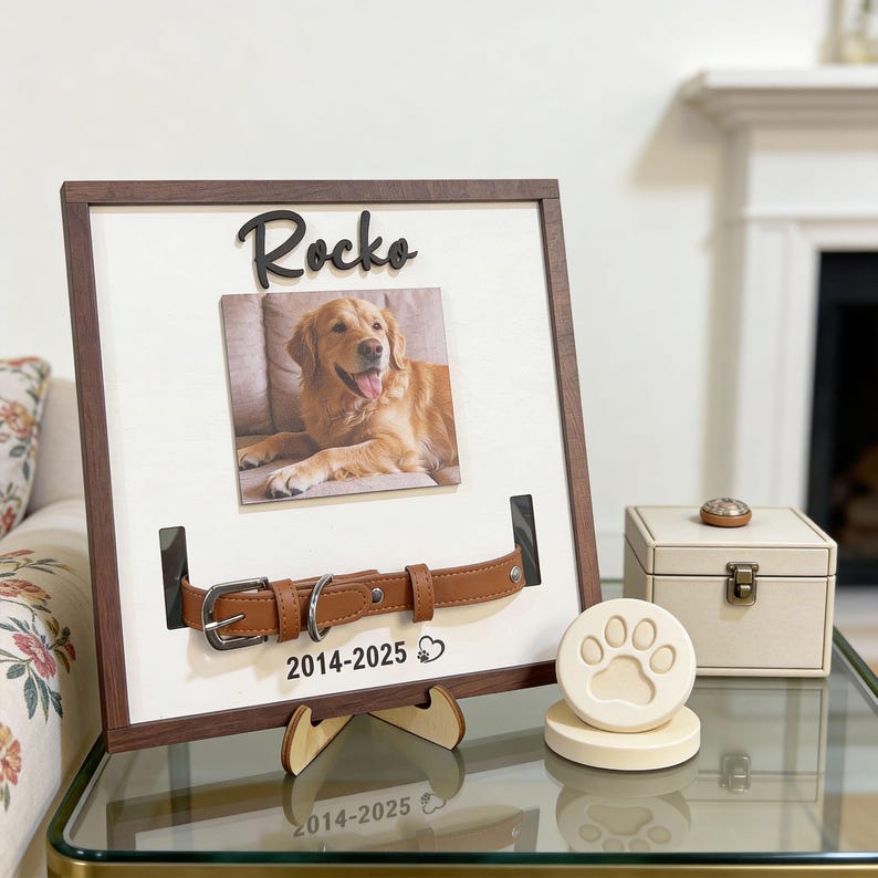 Pu&ograve; includere: Una cornice commemorativa per animali domestici con una foto di un golden retriever, un collare in pelle marrone e il nome "Rocko". Include un sottobicchiere con impronta di zampa e una piccola scatola color crema.