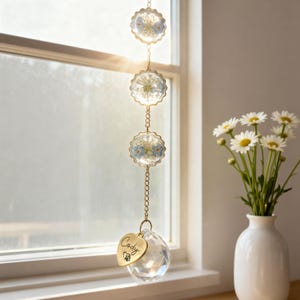 May include: A decorative suncatcher with three circular gold frames holding dried flowers, suspended by a gold chain. A clear crystal ball hangs at the bottom, with a gold heart charm engraved with the name "Cody" and a paw print. The suncatcher is displayed near a window.