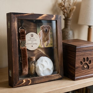 May include: A dark wood shadow box frame containing a brown velvet dog collar, a small glass vial with fur, a white clay paw print, and a photo of a golden retriever. A circular wooden plaque reads, You were my favourite hello and my hardest goodbye.