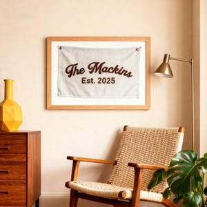 Puede incluir: Un cuadro de pared enmarcado con el texto "The Mackins Est. 2025" en escritura marrón. La obra de arte se muestra sobre una tela de color claro, dentro de un marco de madera natural. La habitación cuenta con una silla de madera y un jarrón amarillo.
