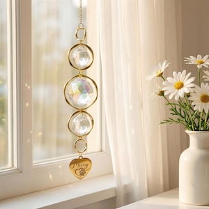 Puede incluir: Un atrapasoles decorativo con tres esferas de cristal y un dije en forma de corazón. El atrapasoles está suspendido por una cadena dorada. El dije de corazón está grabado con el nombre "Miley" y una huella de pata.