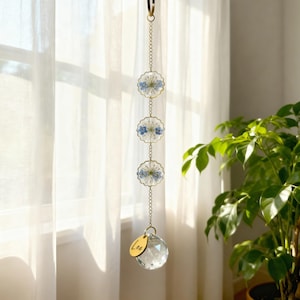 May include: A hanging suncatcher with three circular, gold-toned charms, each containing blue flowers. A clear crystal ball and a gold tag with the name "Lea" hang below. The suncatcher is suspended from a gold chain.
