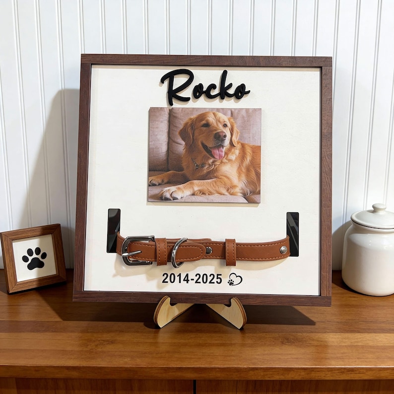 Pu&ograve; includere: Cornice commemorativa per animali domestici in legno con foto di un golden retriever, il nome "Rocko" in corsivo nero e un collare per cani in pelle marrone. La cornice include gli anni "2014-2025" e una zampa.