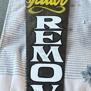 May include: A hand saw with a wooden handle, painted with the words "Tattoo Removal". The design includes a "5¢" sign and the word "Tattoo" in yellow script. The saw blade is dark with white lettering.