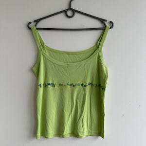 Puede incluir: Una camiseta sin mangas verde lima con tirantes finos, colgada de una percha negra. La parte superior presenta un diseño floral horizontal en azul y amarillo en el pecho. La tela parece ser un material suave y ligero.