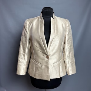 Könnte beinhalten: Hellbeige Blazer mit Reverskragen und Einzelknopfverschluss. Der Blazer hat zwei Vordertaschen und wird auf einer schwarzen Schaufensterpuppe vor grauem Hintergrund präsentiert. Der Stoff hat einen dezenten Glanz.