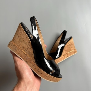 Puede incluir: Par de sandalias de cuña negras de charol con punta abierta y tacones de corcho. Las sandalias tienen una correa trasera y un ribete dorado a lo largo de la suela. Los tacones de cuña miden aproximadamente 10 cm de alto.